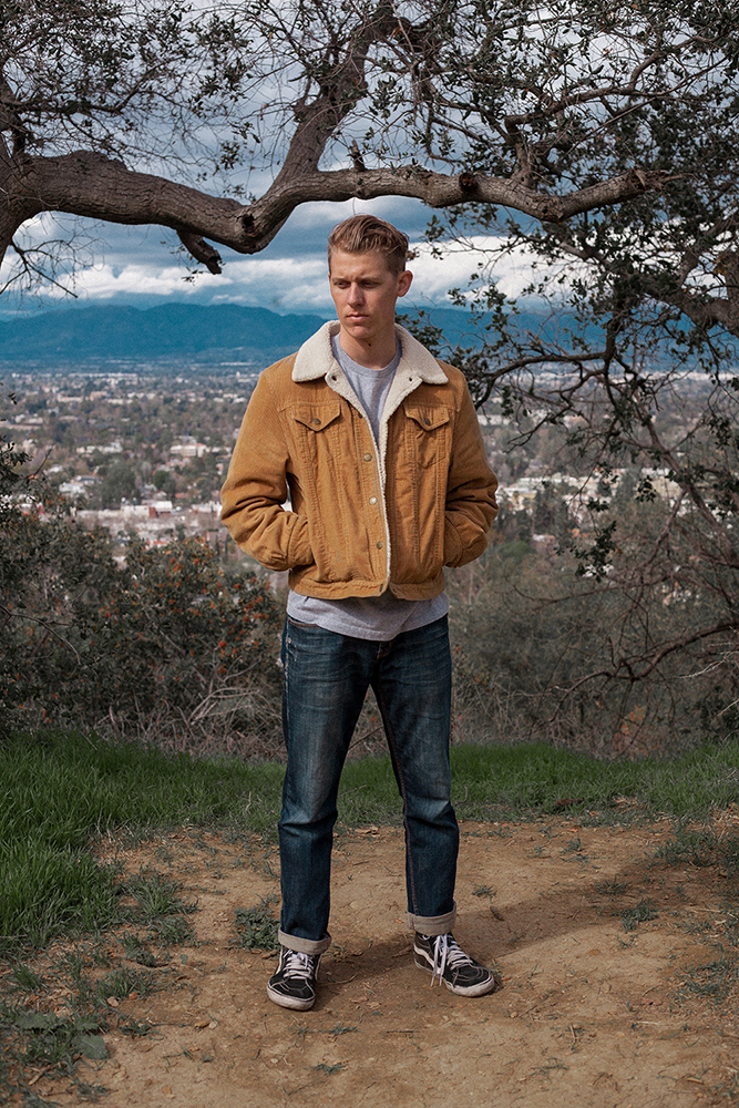 Man standing in the woods, town behind him wearing a brown jacket.